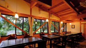 A dining area with wooden tables and chairs, large windows with curtains, and hanging pendant lights.
