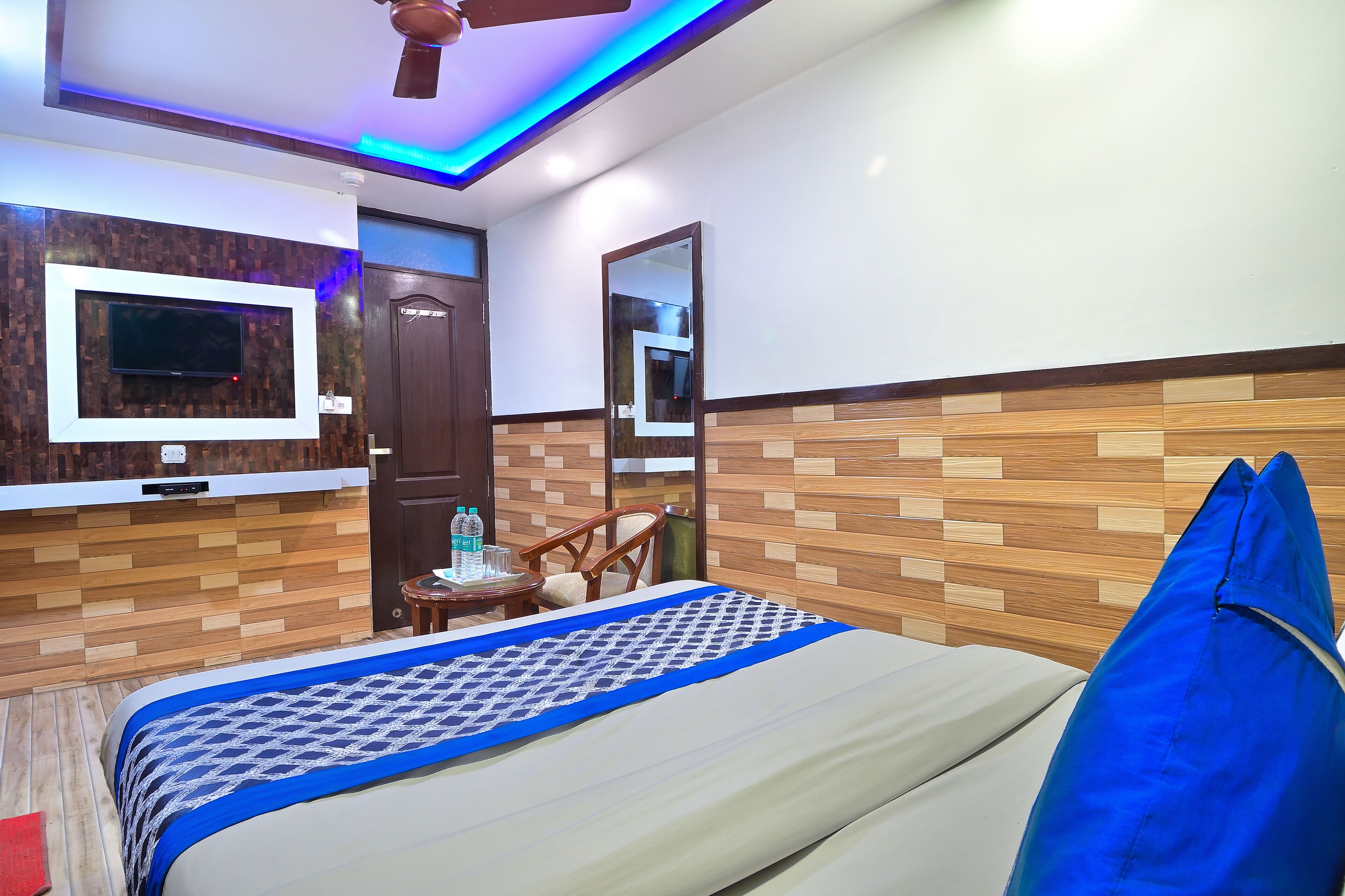Una camera d'albergo con un letto, una TV a muro, un pannello decorativo in legno, una ventola a soffitto e un piccolo tavolo con una sedia.