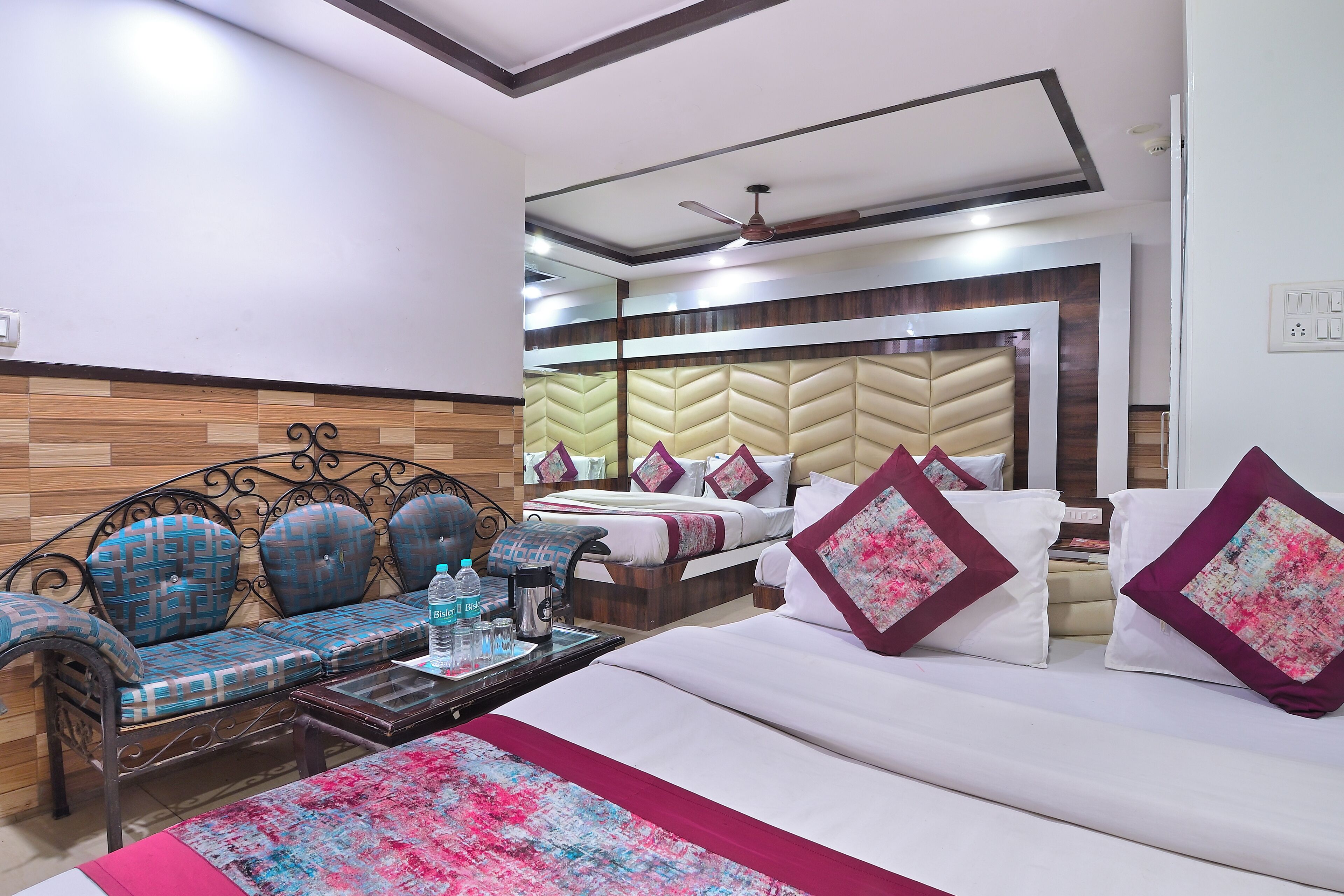 A hotel room with a sofa, a bed with decorative pillows, a coffee table with a water bottle, and a ceiling fan.