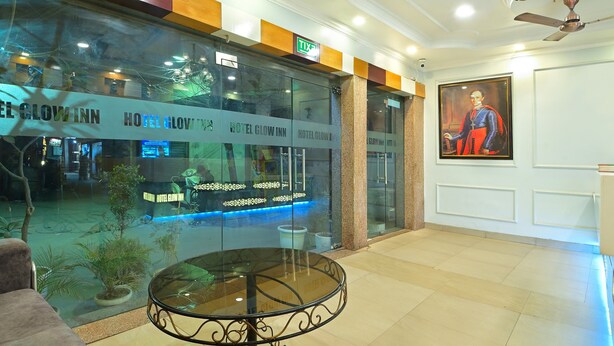 A hotel lobby with a reception desk, a glass wall with the hotel name, a chandelier, and a painting on the wall. - Hotel Glow Inn (New Delhi)