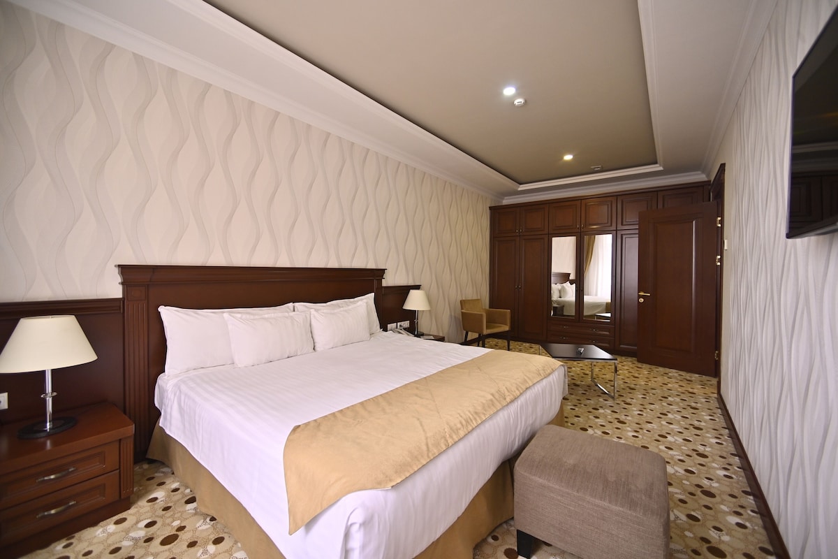 A hotel room with a large bed, a bedside table with a lamp, a chair, and a small table.
