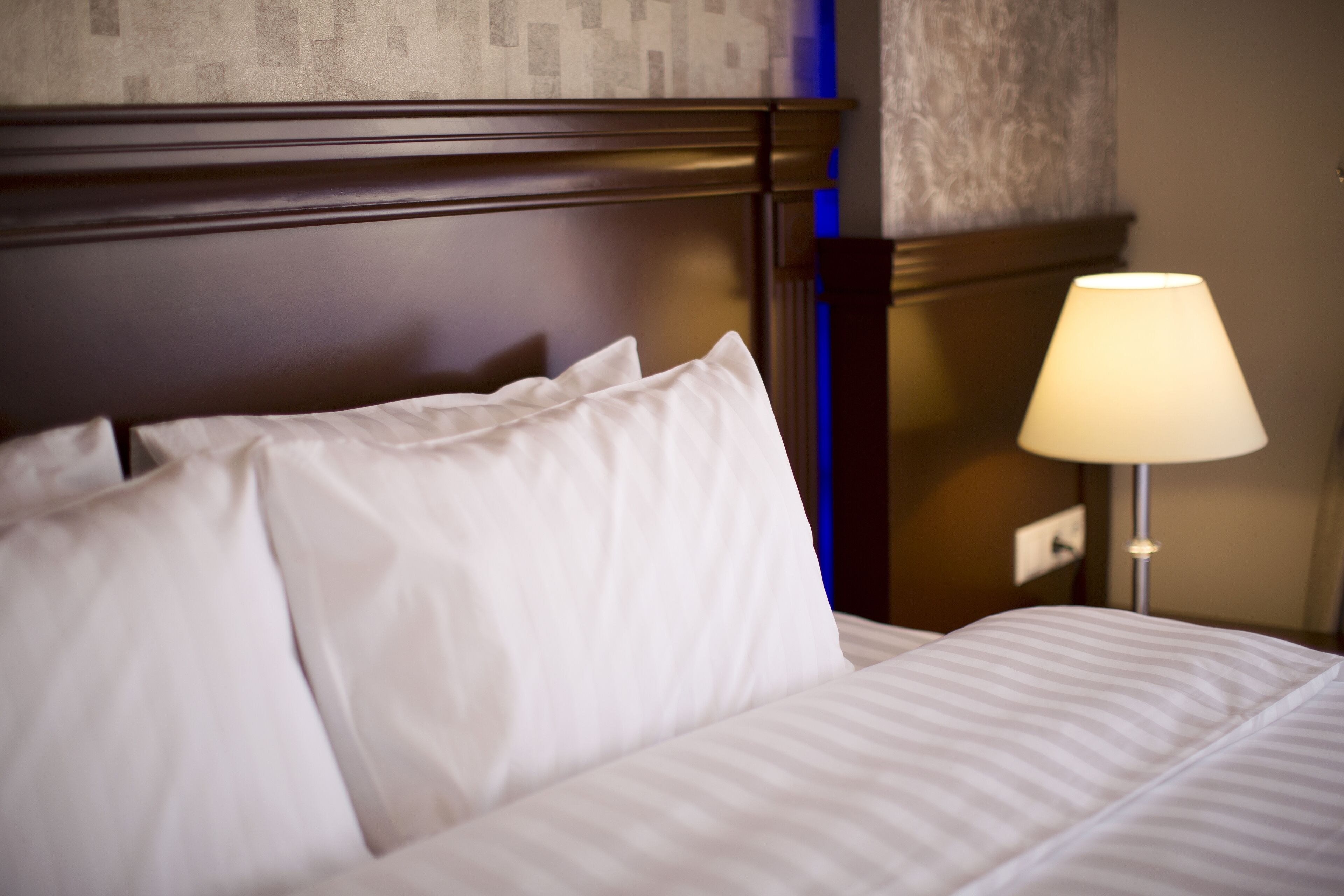 Una cama bien hecha con sábanas blancas y un cabecero de madera. Una lámpara de noche está encendida, proyectando una luz suave.
