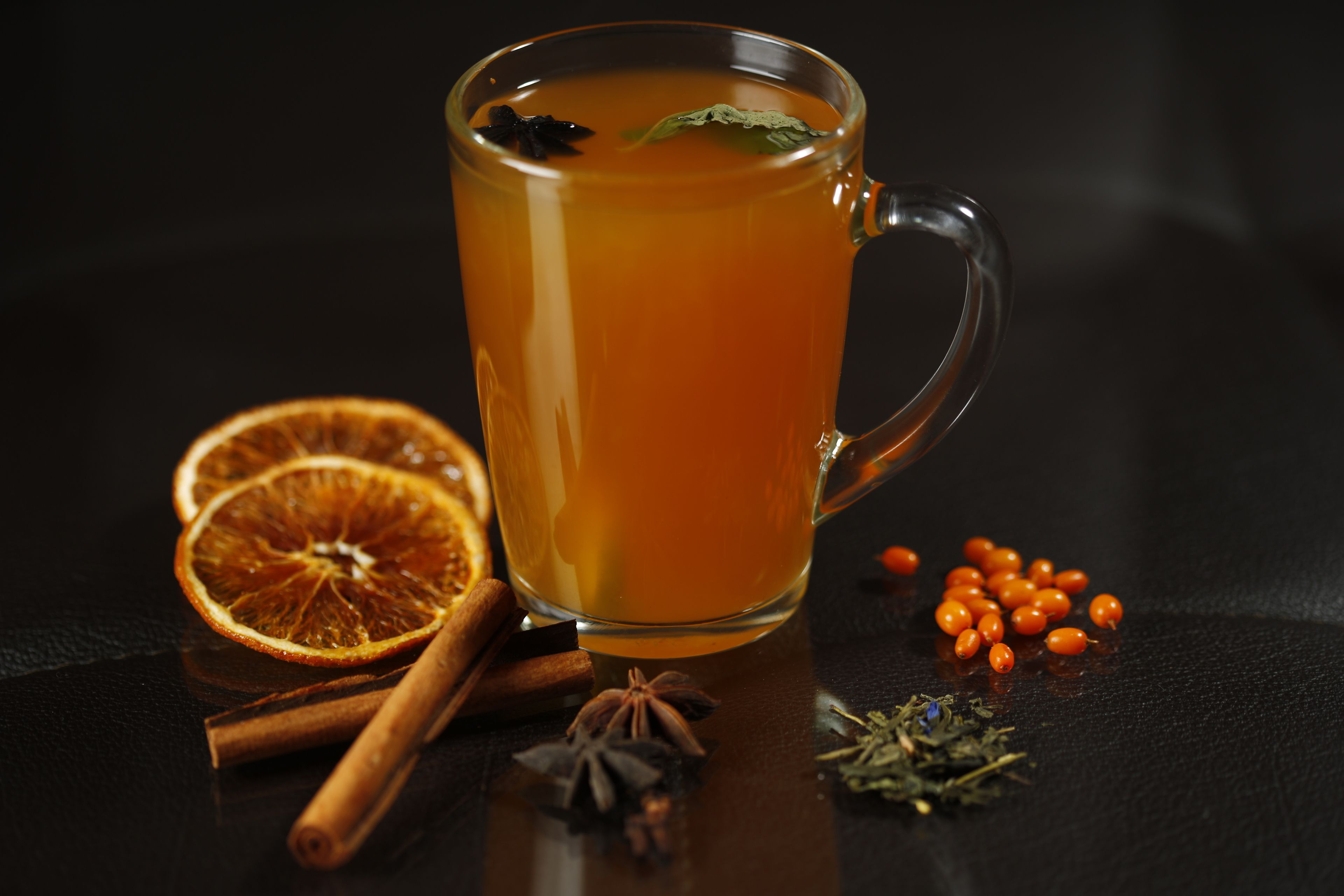 Una taza de vidrio transparente llena de una bebida, acompañada de rodajas de naranja deshidratada, ramas de canela, anís estrellado y pequeñas bayas de naranja sobre una superficie oscura.