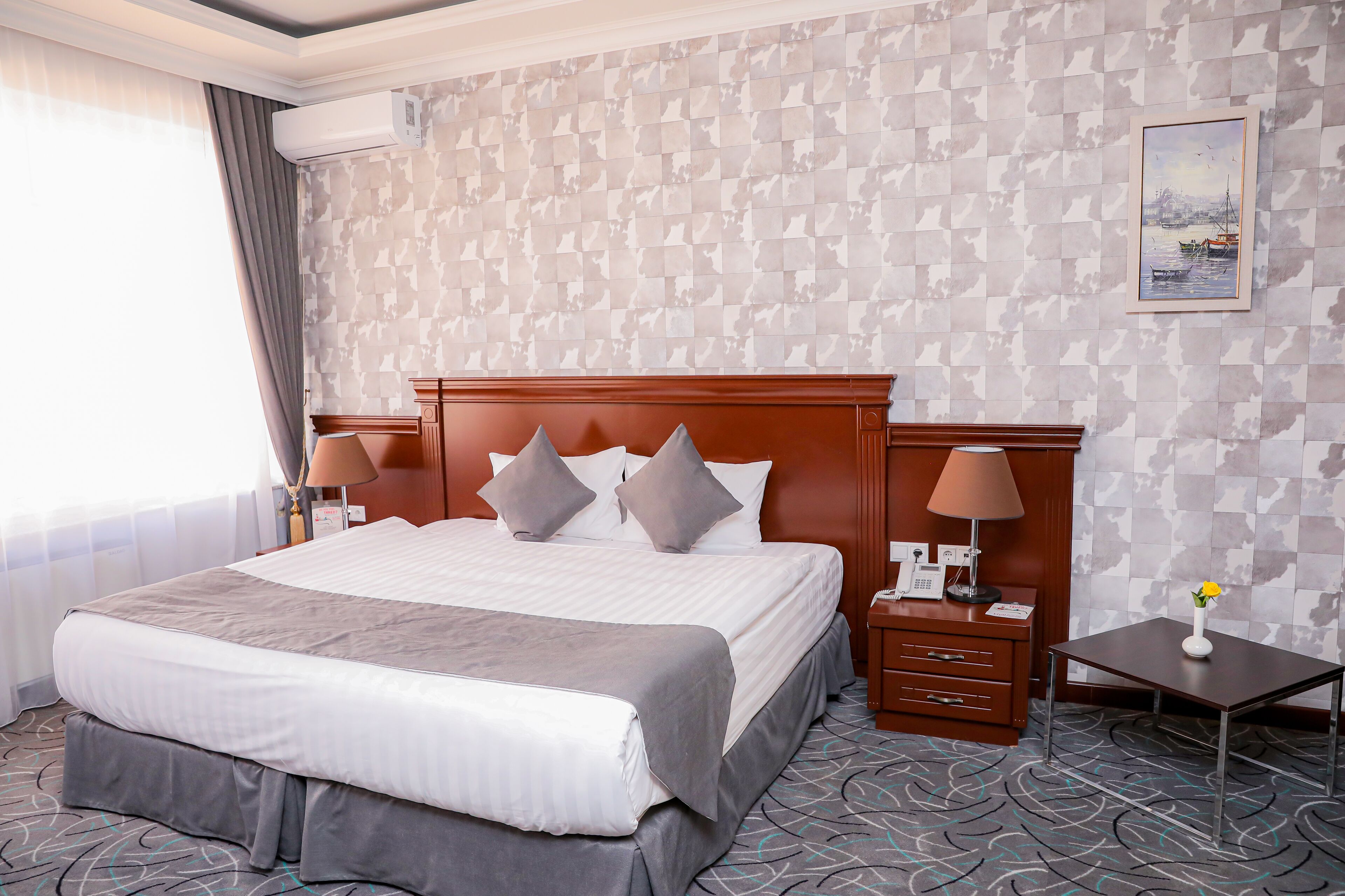 A hotel room with a large bed, a wooden nightstand, a desk, and a wall-mounted air conditioner.