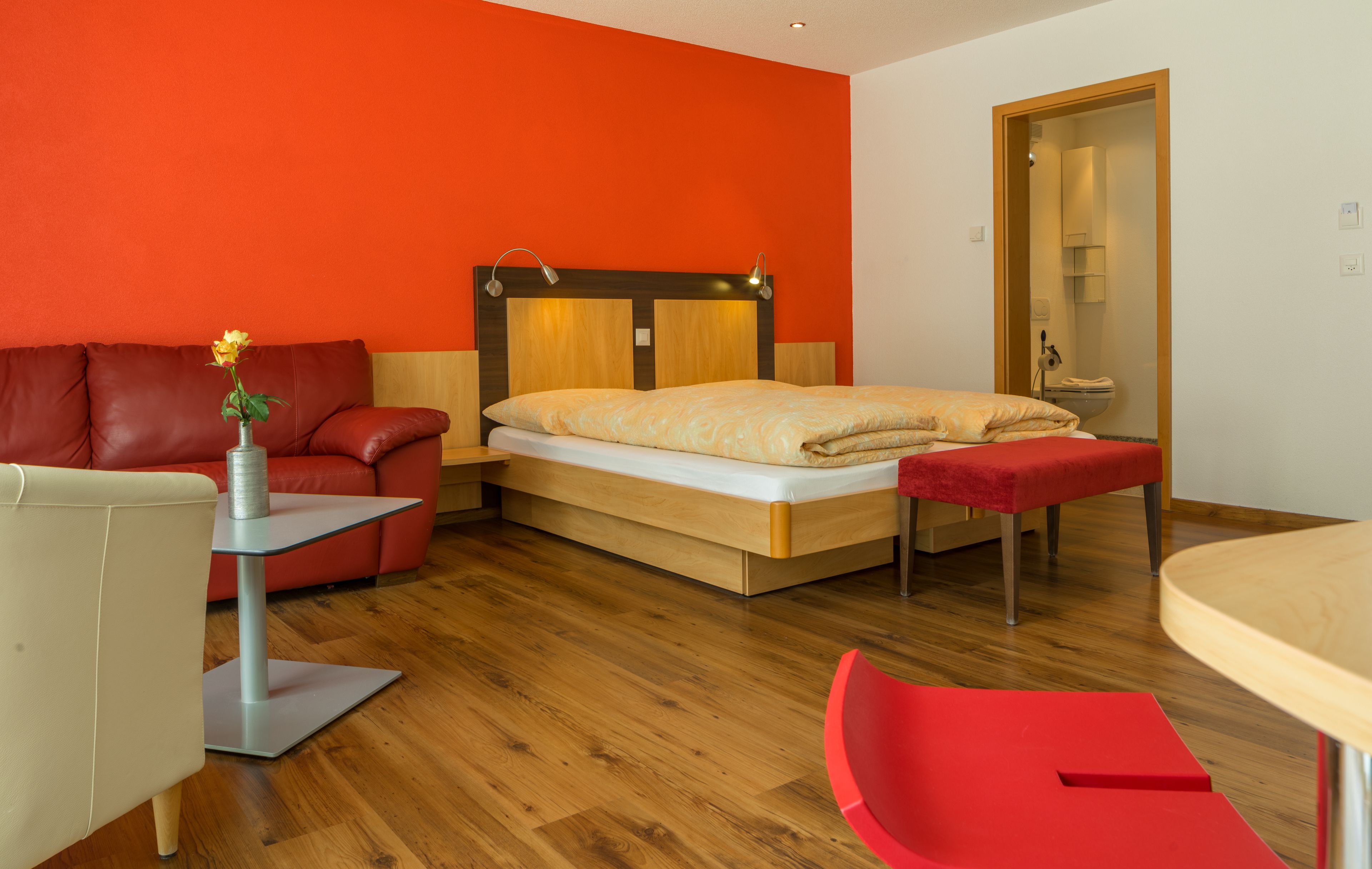 Une chambre d’hôtel moderne avec un plancher en bois, un lit doté d’une tête de lit en bois, un canapé rouge, une petite table et une chaise rouge.