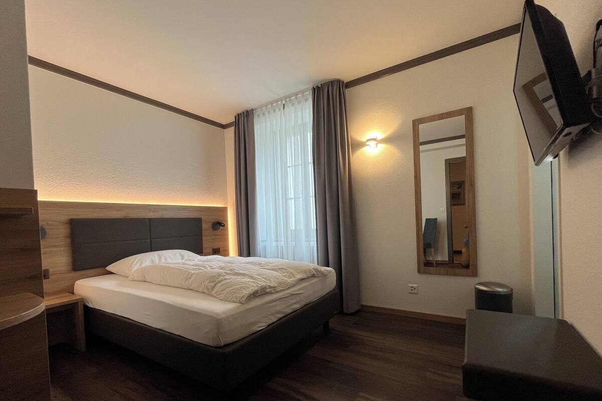 Una habitación de hotel moderna con una cama, una mesita de noche, un espejo y un televisor fijado a la pared.