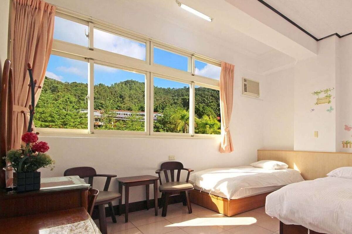 A room with two beds, a desk with chairs, a window with curtains, and a view of greenery outside.