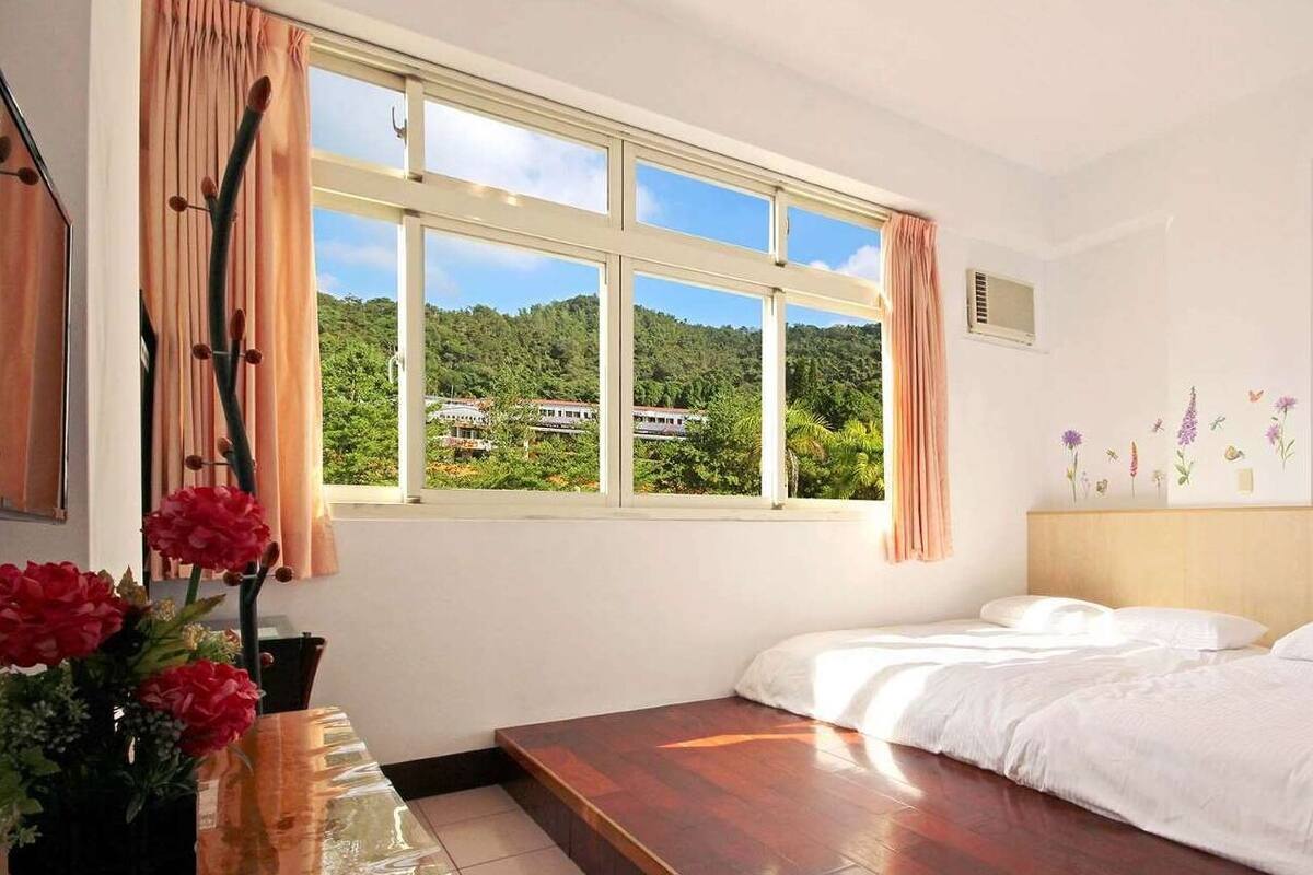 A bedroom with a large window, a bed, a bedside table with flowers, and a wall with floral decals.