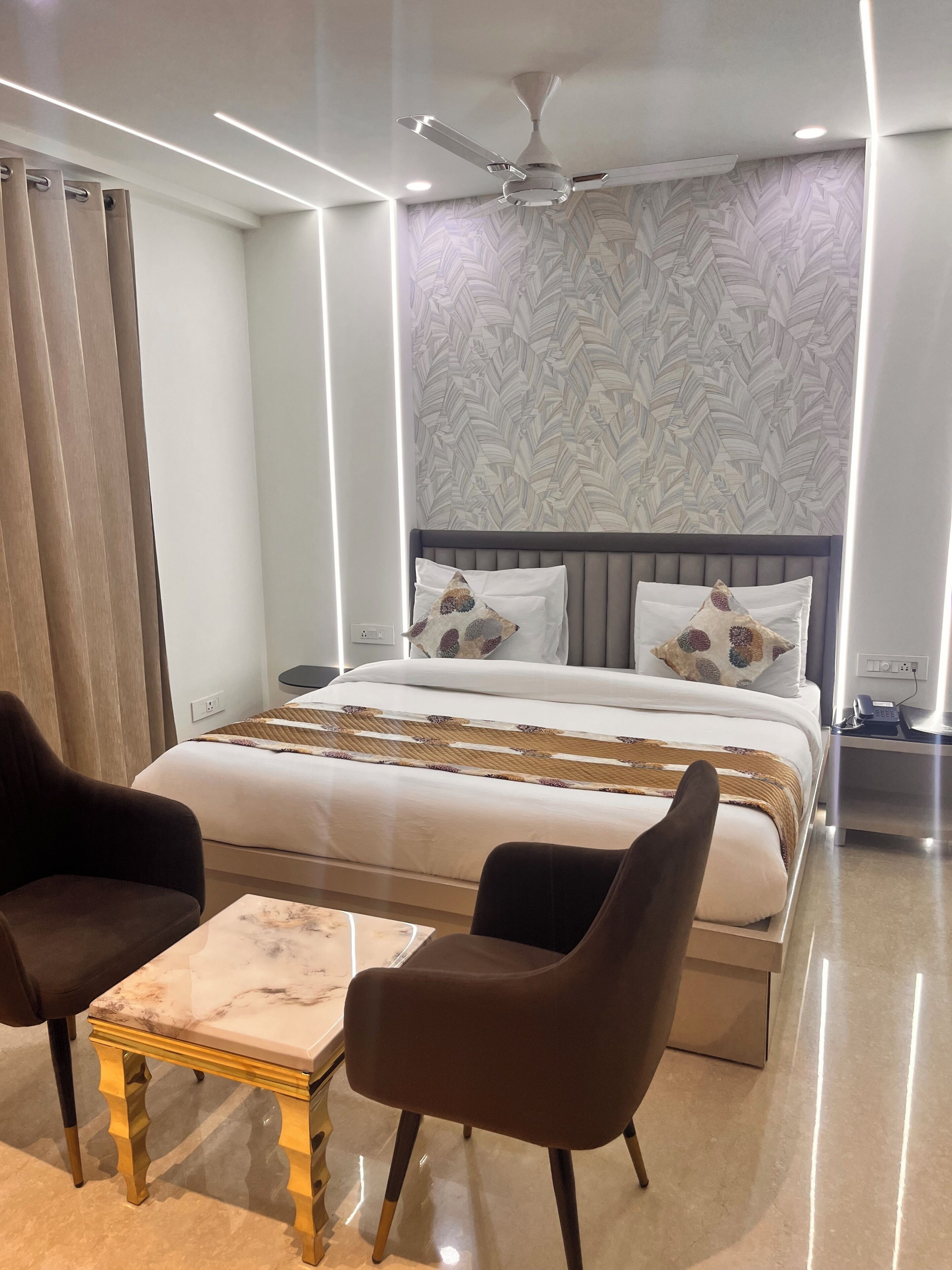 A modern hotel room with a large bed, two armchairs, a small table, and a ceiling fan.