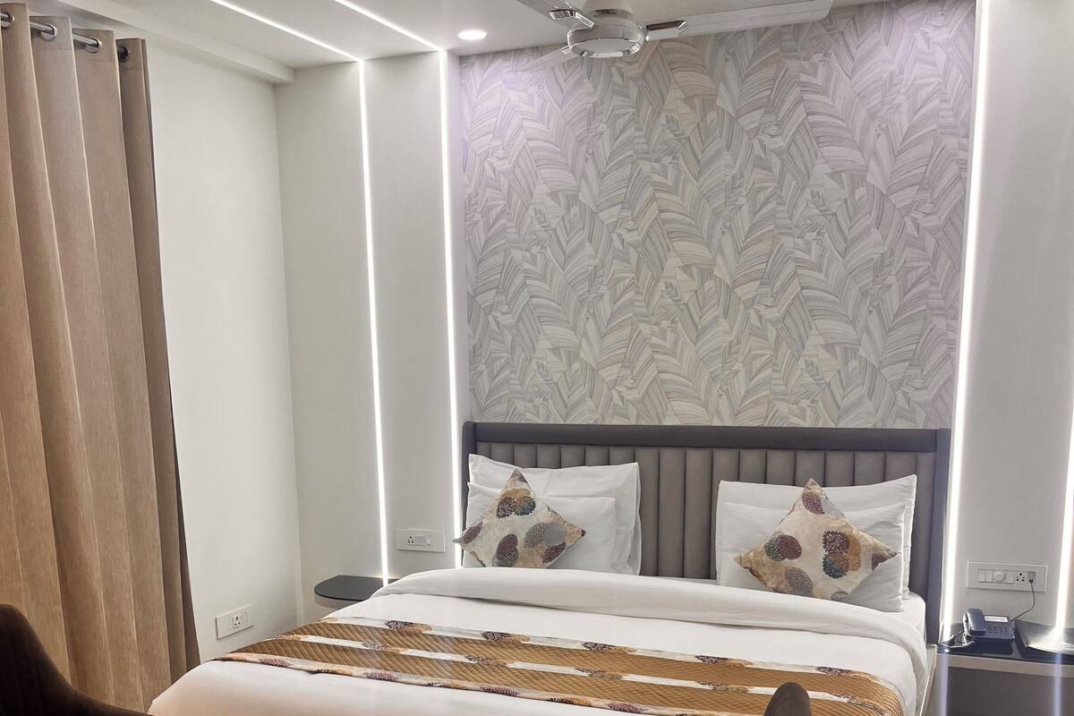 A modern hotel room with a large bed, two armchairs, a small table, a ceiling fan, and a patterned wall.