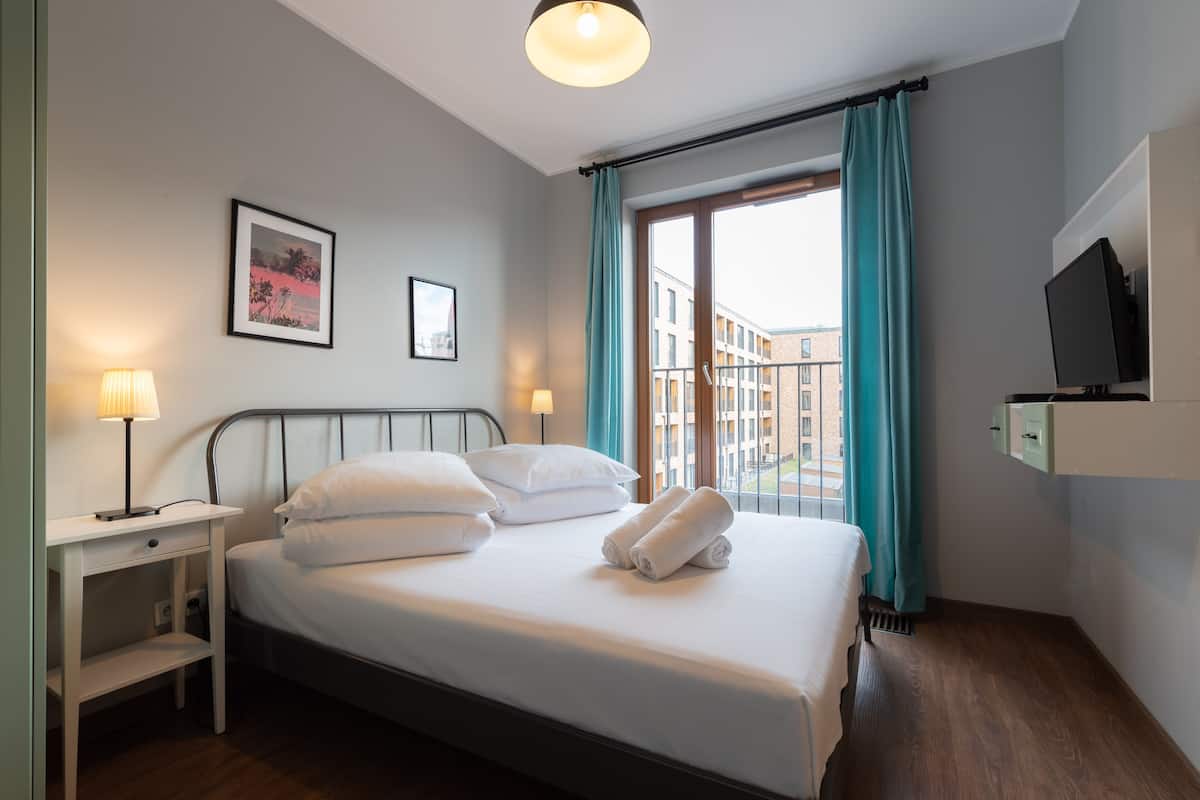 Een opgemaakt bed met kussens en handdoeken, een nachtkastje met een lamp, een lijst met een foto aan de muur en een raam met gordijnen.