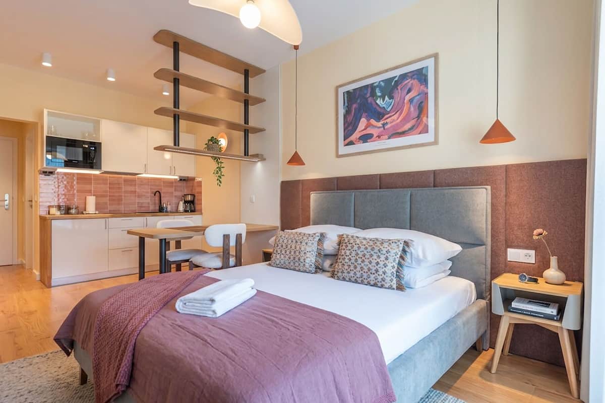 Een moderne slaapkamer met een groot bed, nachtkastje, bijzetmeubel, hoofdeinde en uitzicht op een keukenhoek.