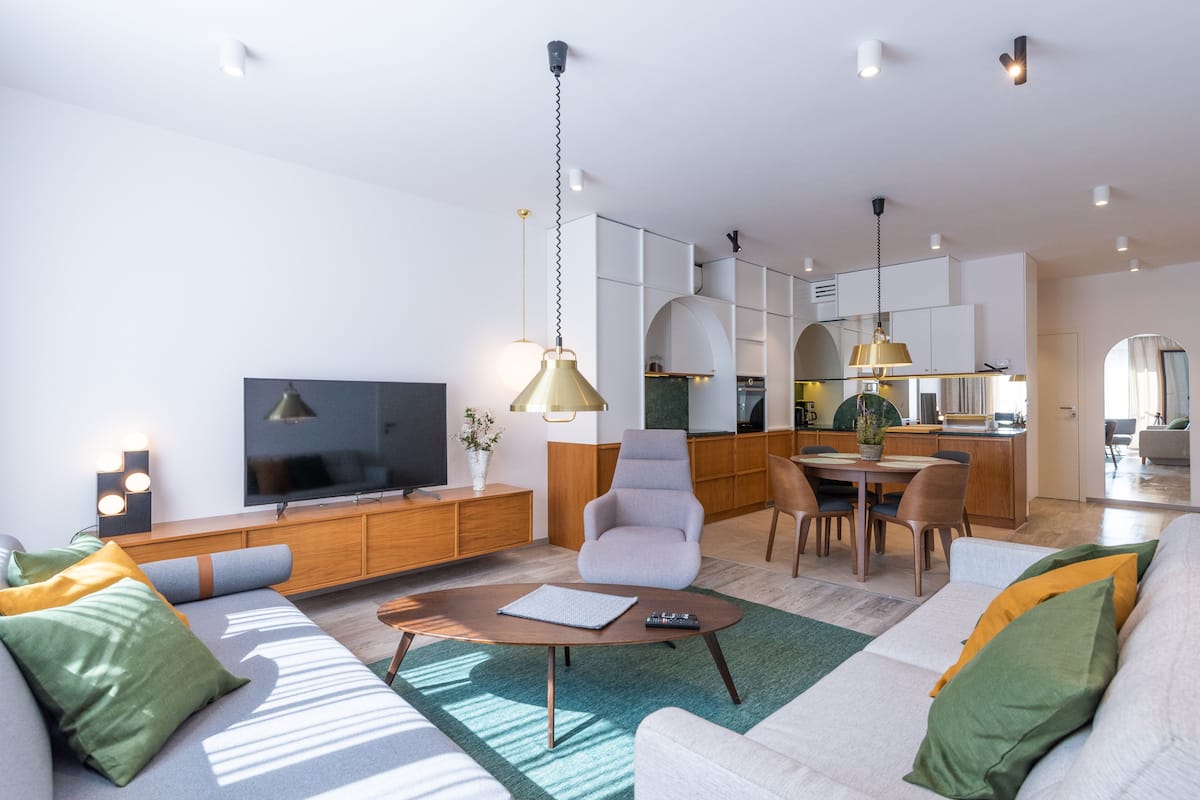Een moderne woonkamer met een bank, salontafel en televisie. Er is een eethoek met stoelen en een tafel. De keuken is zichtbaar op de achtergrond.