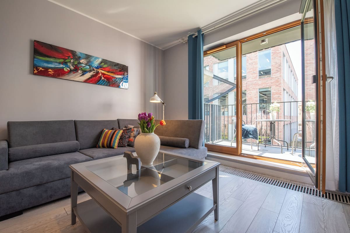 Een moderne woonkamer met een grijze bank, een glazen salontafel en een kleurrijk abstract schilderij aan de muur.