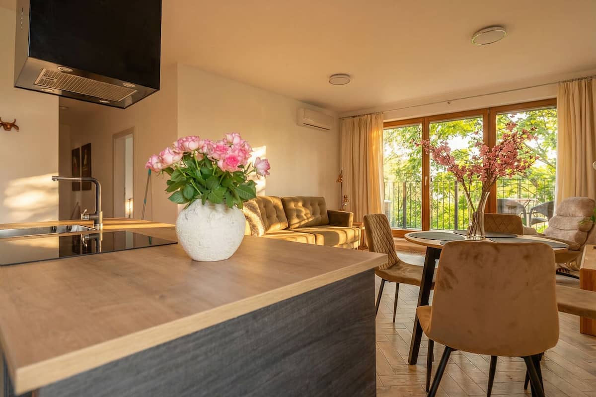 Een moderne keuken met een ingebouwde kookplaat, een eethoek met een ronde tafel en stoelen, een bank en een groot raam met uitzicht op groen.