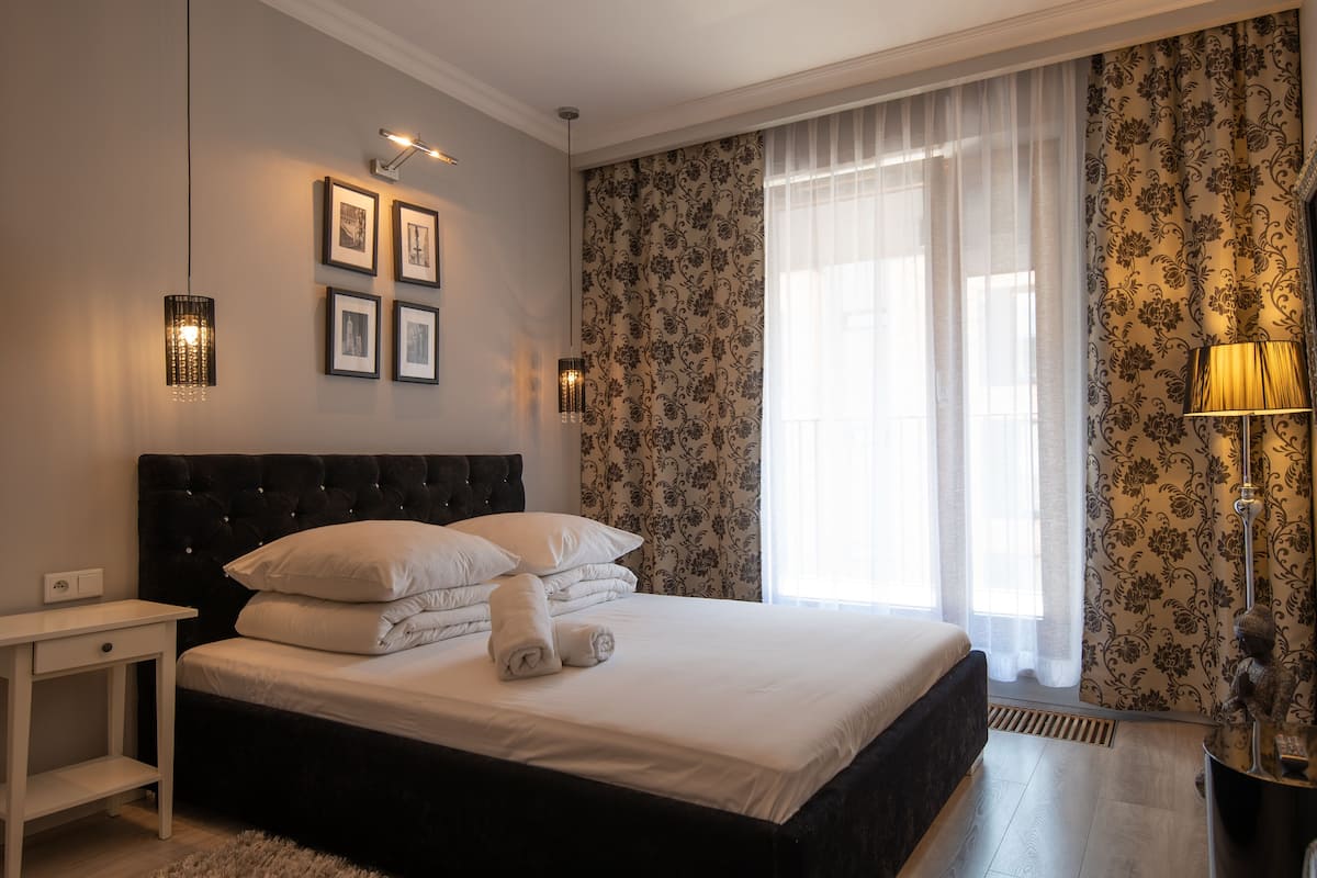 Een net opgemaakt bed met witte lakens en kussens, een nachtkastje met een lamp, en ingelijste foto's aan de muur.