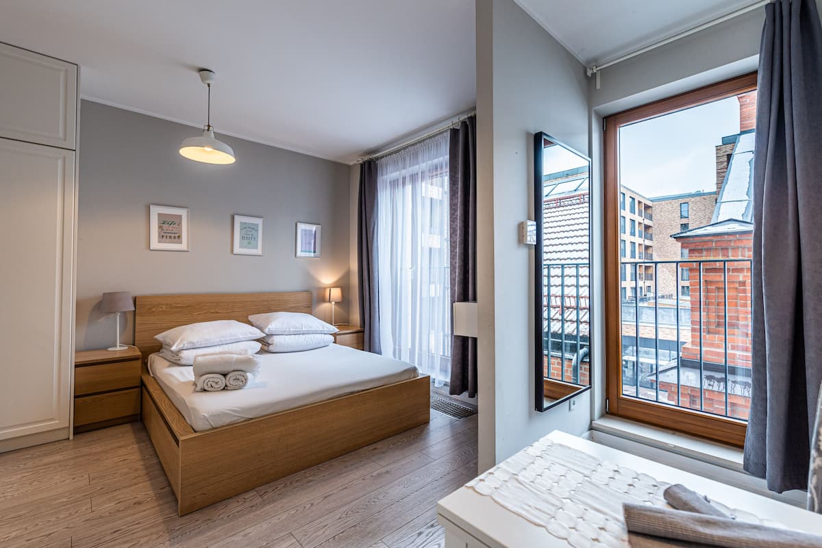 Een moderne hotelkamer met een houten bed, bedlampen, een groot raam met uitzicht op gebouwen, en lijstwerken aan de muur.