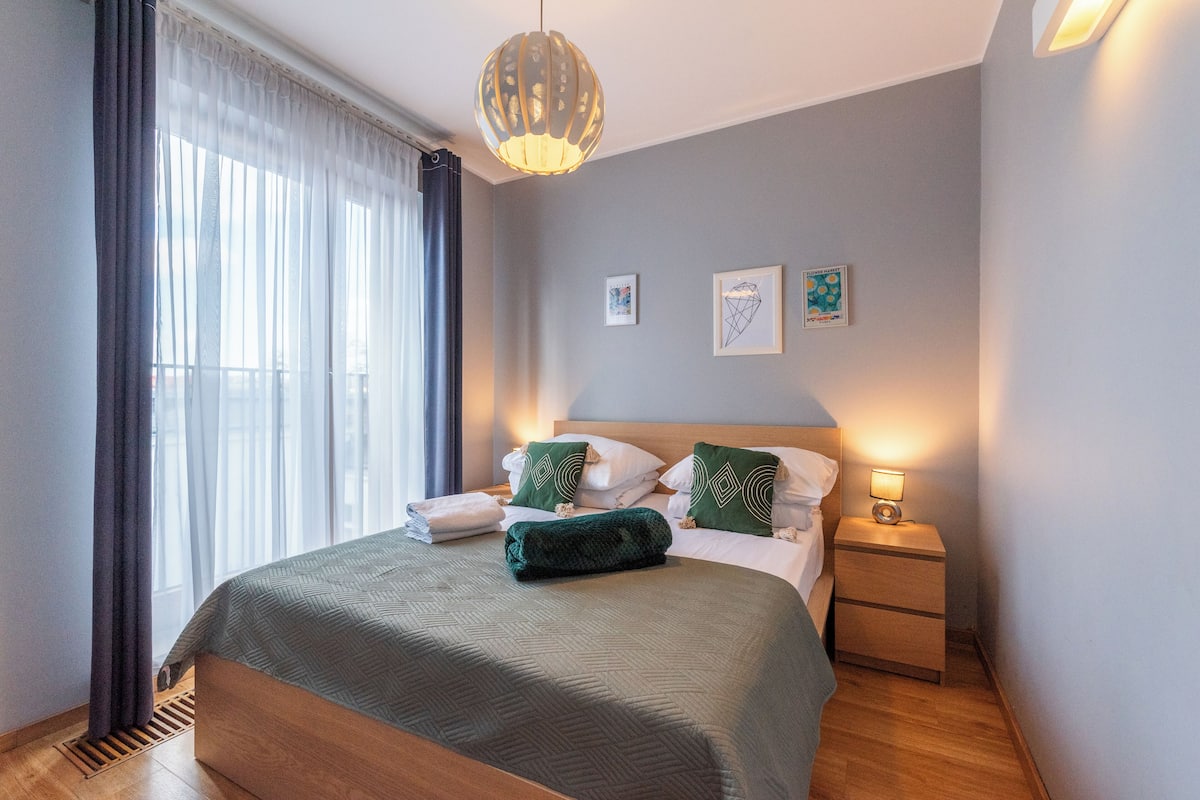 Een net opgemaakt bed met een groene deken en kussens, een nachtkastje met een lamp en een raam met dunne gordijnen.