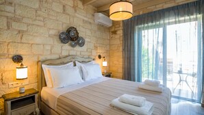 A bedroom with a stone wall, a bed with white linens, two bedside lamps, and a view of the outdoors through a large window. - Villa Sirocos, Private eco pool, near the beach (Chania)