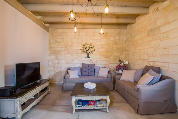 A cozy living room with a stone wall, a flat-screen TV on a wooden stand, a grey sofa, a coffee table, and a ceiling with exposed beams and hanging light fixtures. - Villa Sirocos, Private pool, near the beach (Chania)