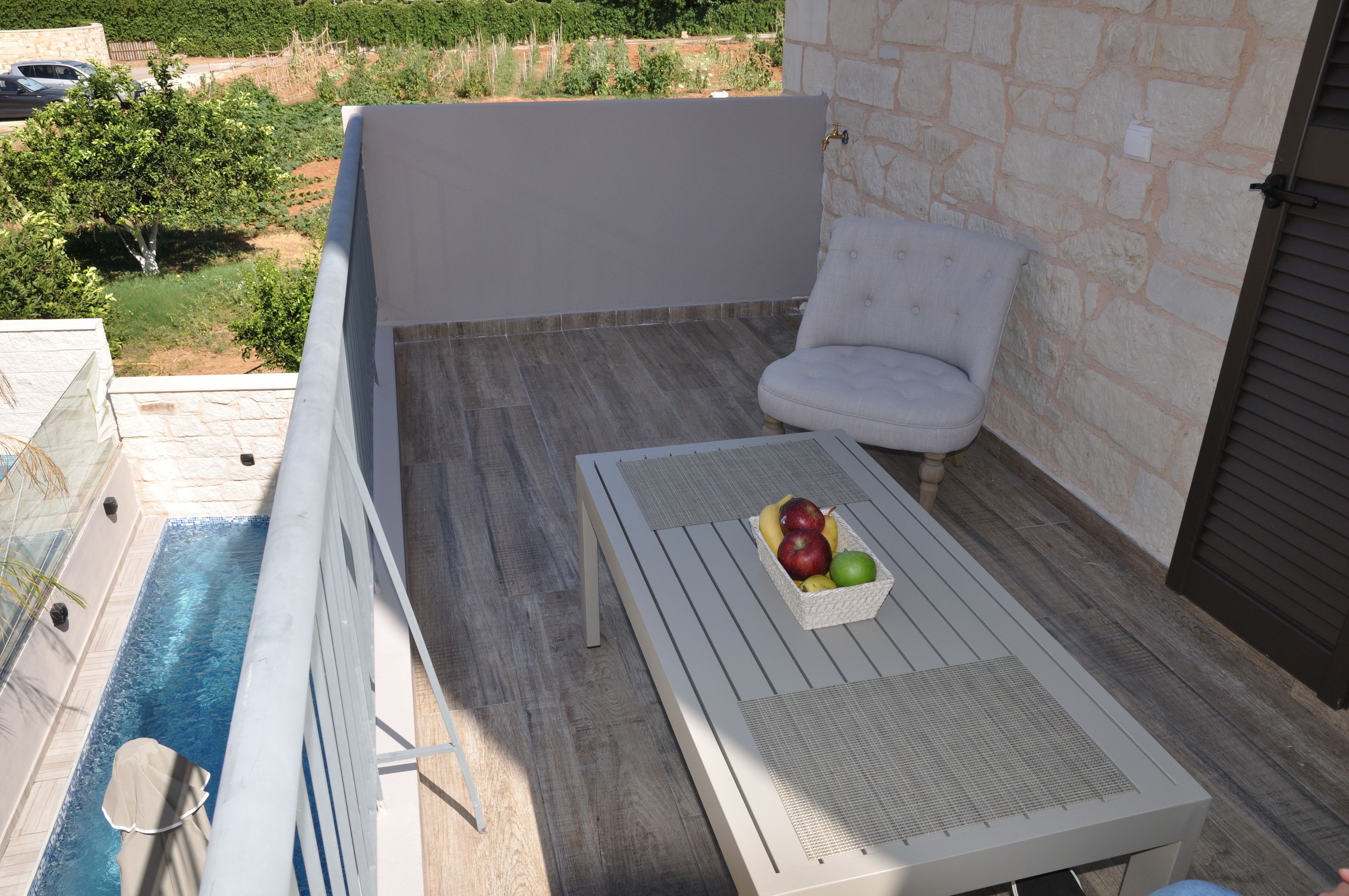 A balcony with a white chair, a table with a fruit basket, and a pool.