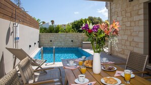 A poolside breakfast setup with lounge chairs, a table with plates of food, drinks, and a vase of flowers. - Villa Sirocos, Private eco pool, near the beach (Chania)