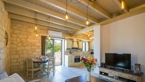 A spacious living area with a stone wall, wooden ceiling beams, a dining table with chairs, a sofa, a TV on a wooden stand, and a kitchen in the background. - Villa Sirocos, Private eco pool, near the beach (Chania)