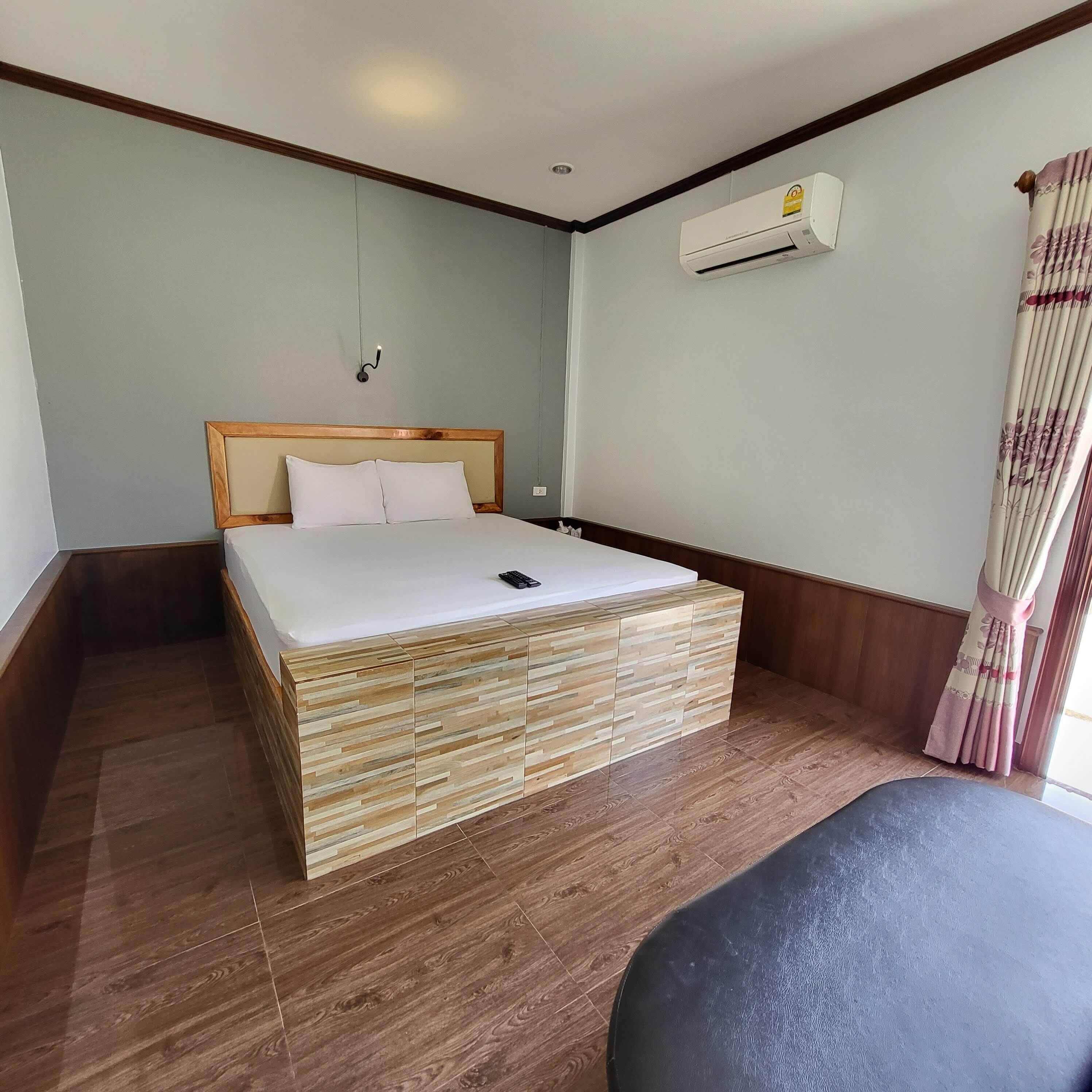 Een hotelkamer met een houten bed, een klein nachtkastje, een wandairconditioner en een raam met gordijnen.