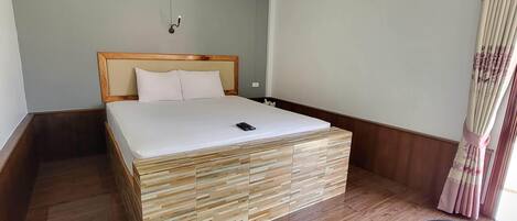 A hotel room with a wooden bed, a small bedside table, a wall-mounted air conditioner, and a window with curtains.