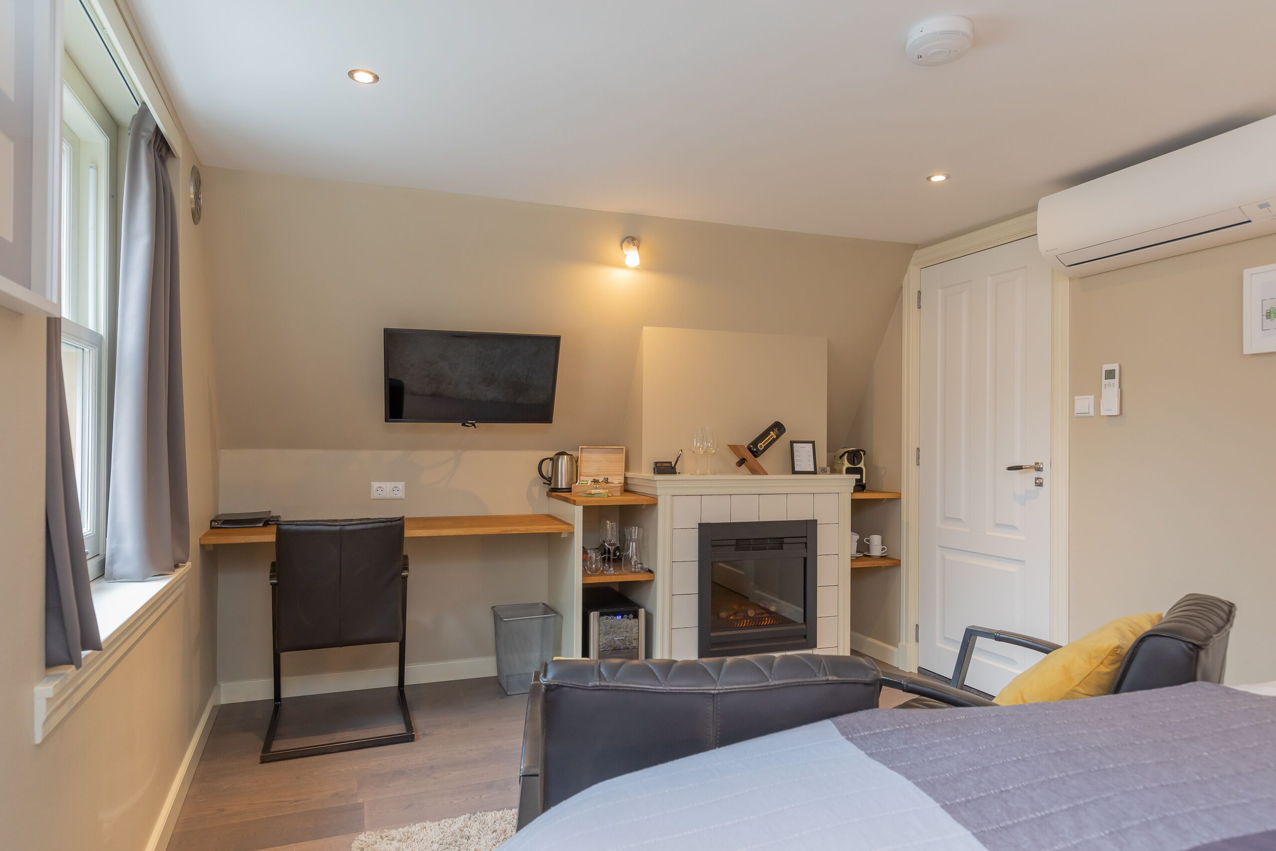 A compact hotel room with a bed, a desk, a chair, a fireplace, and a television.