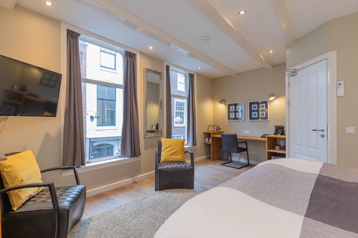 A modern hotel room with a large bed, a desk, a chair, a TV, and large windows with curtains.