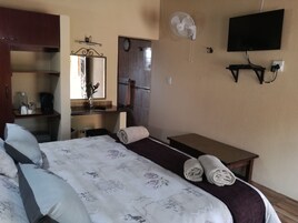 Una camera d'albergo con un letto, un comodino, uno specchio, una TV, un ventilatore e un bagno visibile attraverso una porta aperta.