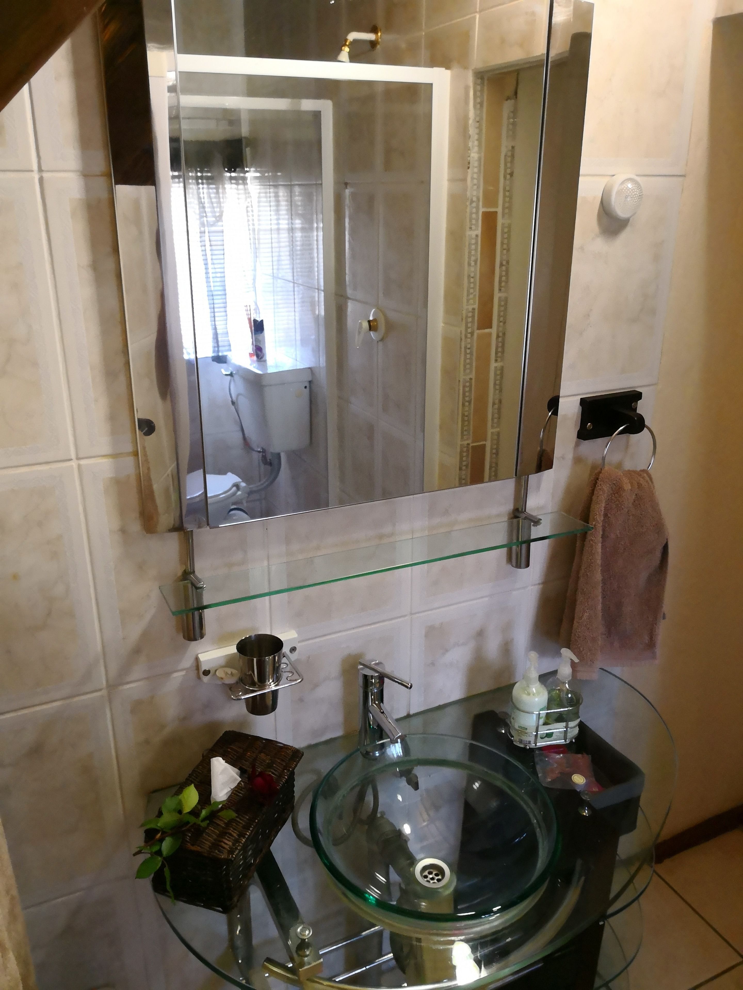 Un baño con lavamanos de vidrio, un espejo, un perchero para toallas y un estante de vidrio con artículos de tocador.
