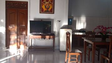 A room with a wooden door, a flat-screen TV on a wooden stand, a refrigerator, a dining table with chairs, and a vase with flowers.