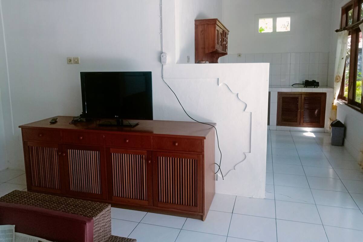 A living room with a TV on a wooden cabinet, a small table, and a kitchen area with cabinets and a window.