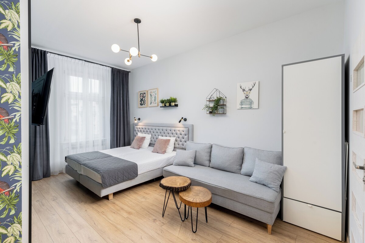 Et moderne soveværelse med en seng, en grå sofa, et lille rundt bord, et fjernsyn og et køleskab.