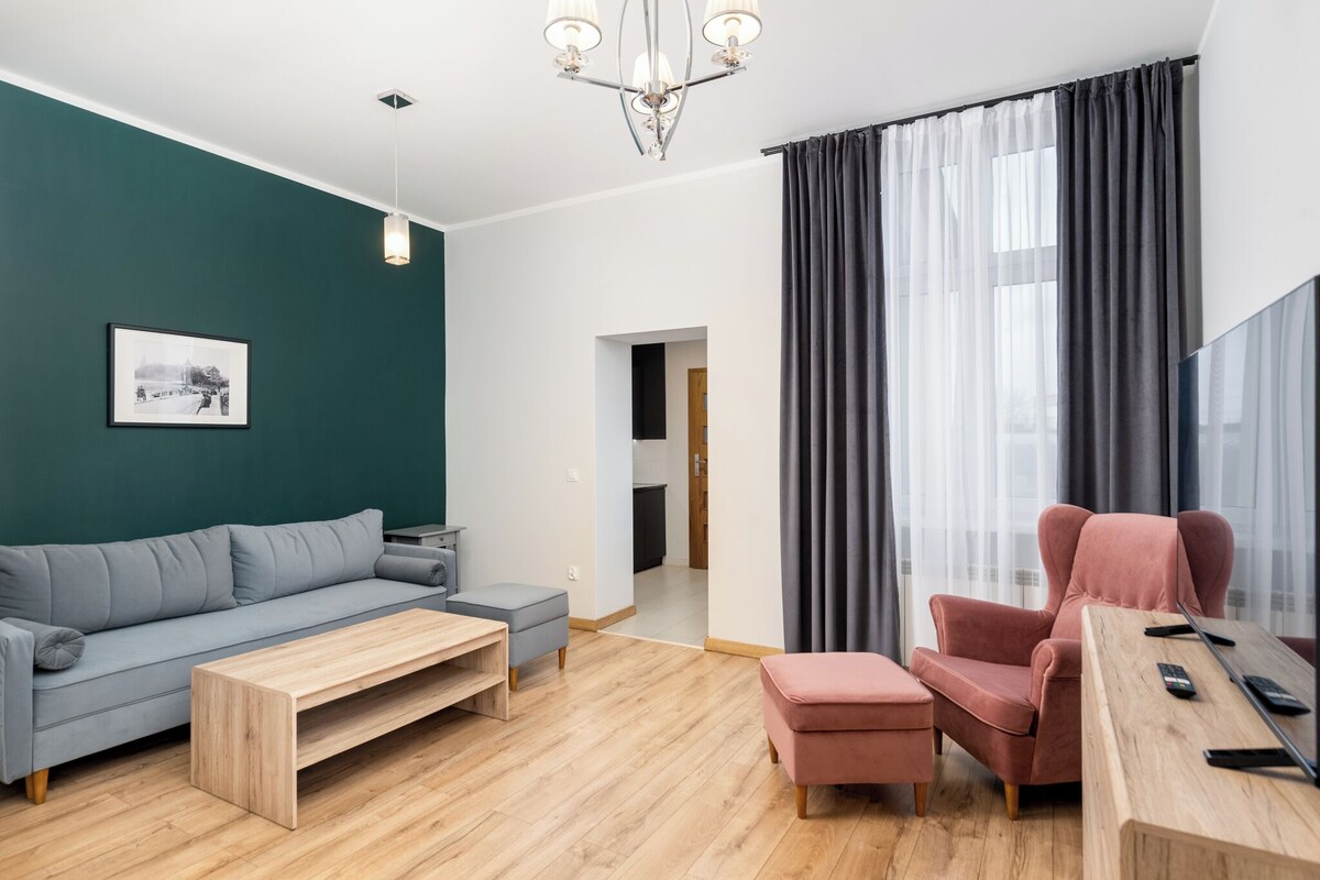 En moderne stue med sofa, skammel, sofabord og en lyserød lænestol. Der er trægulv, en lysekrone og et indrammet billede på væggen.