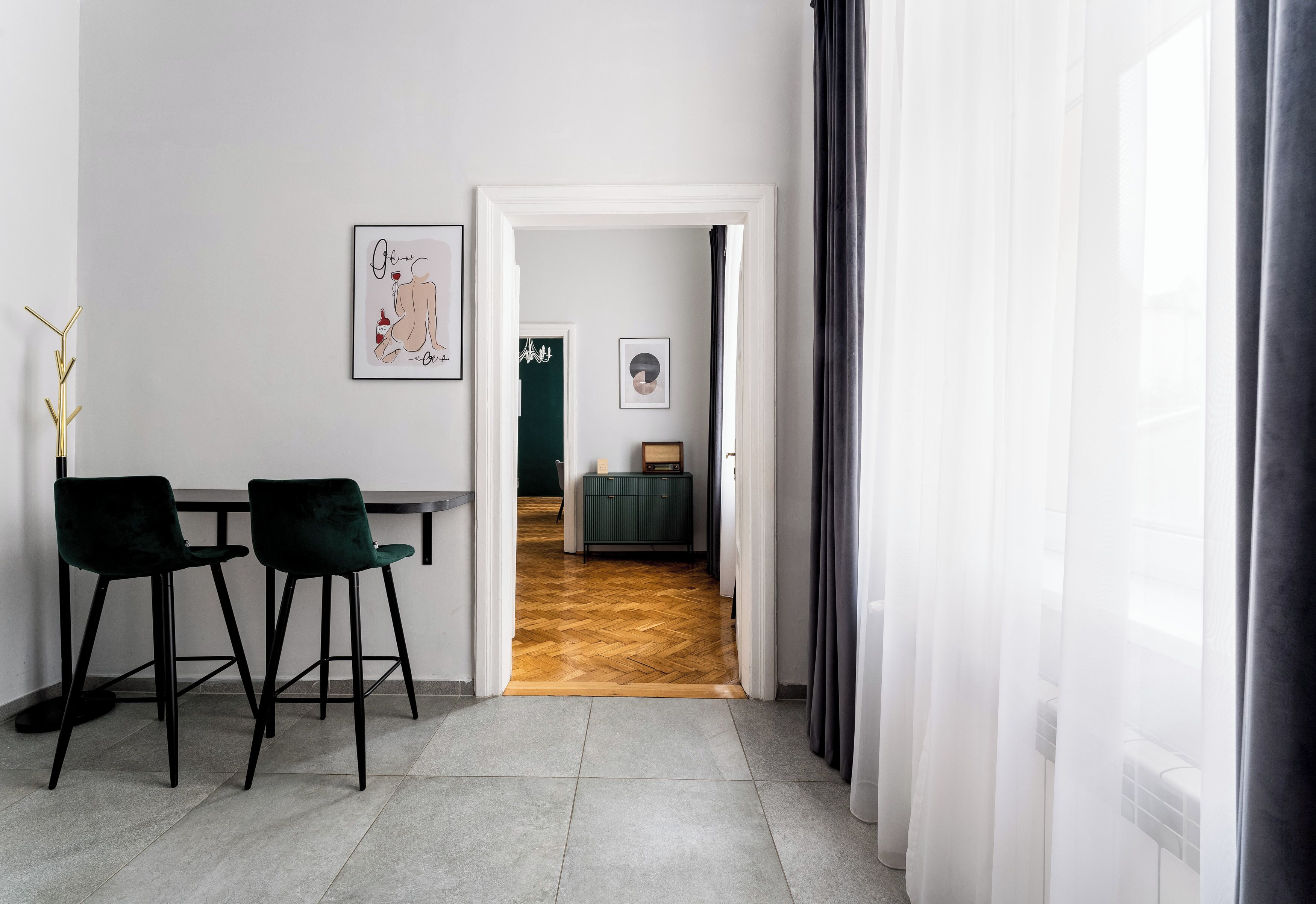 A modern interior with a dining area featuring two chairs and a table, a hallway leading to a room with a green door, and a window with sheer curtains.