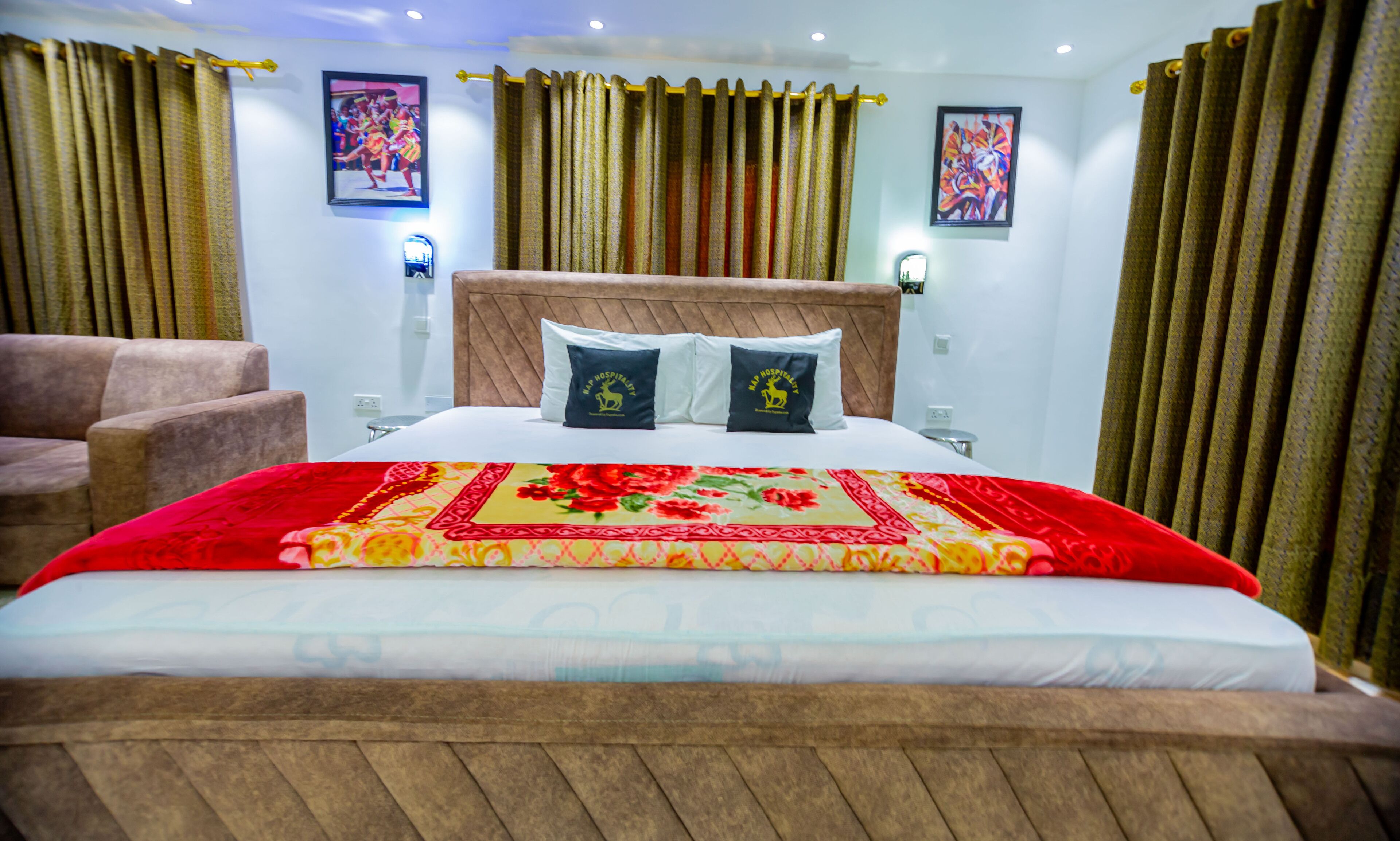 A hotel room with a large bed, two pillows with a logo, a wooden headboard, curtains, and two framed pictures on the walls.
