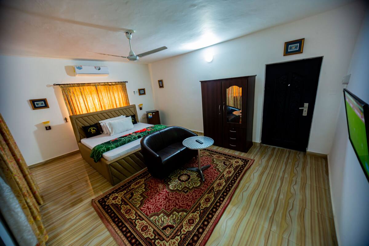 A bedroom with a bed, a black sofa, a small table, a ceiling fan, an air conditioner, a door, and a patterned rug.