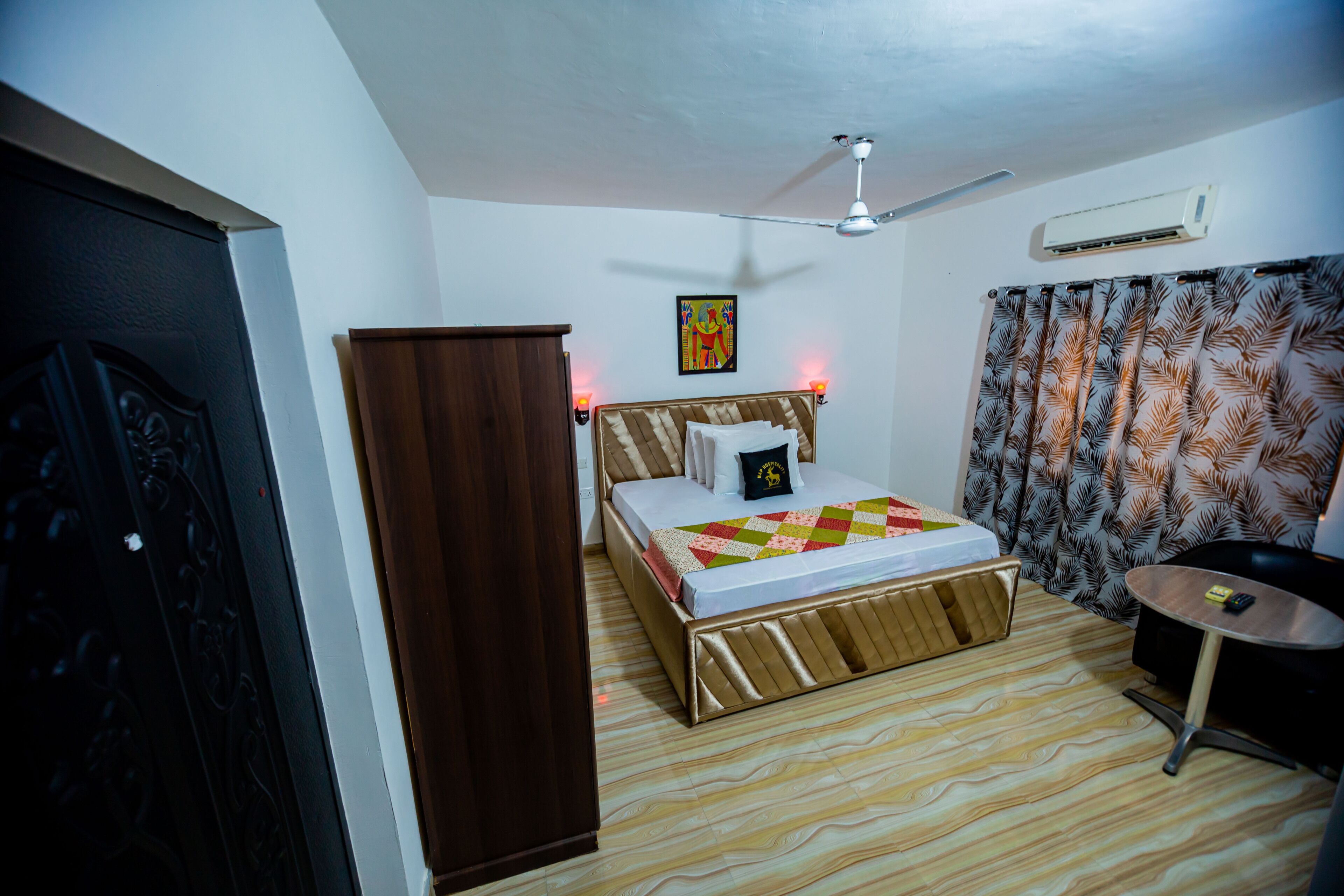 A bedroom with a bed, a wardrobe, a ceiling fan, an air conditioner, and a small table.