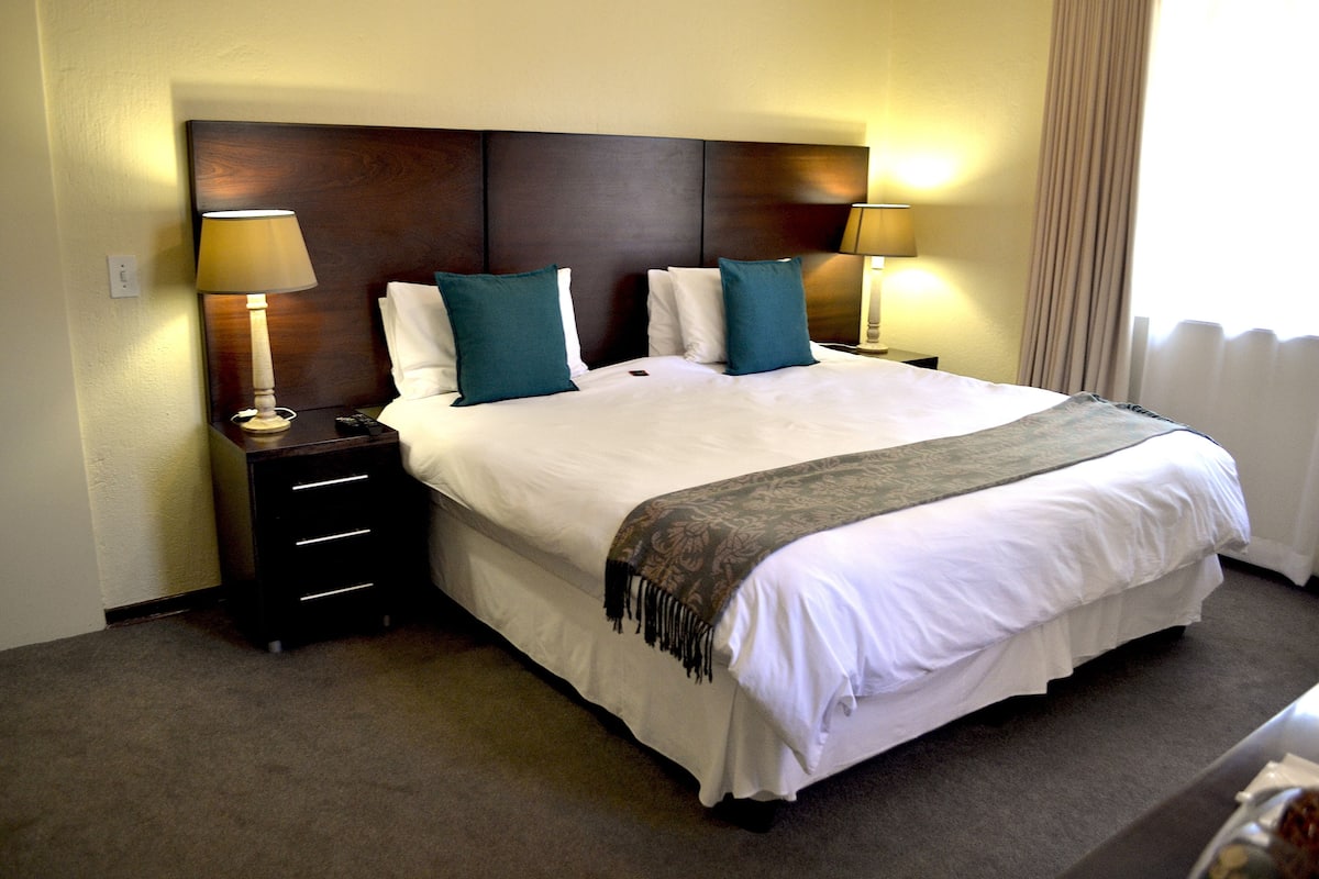 Une chambre d’hôtel avec un grand lit, deux tables de chevet avec des lampes, une tête de lit et une fenêtre avec des rideaux.