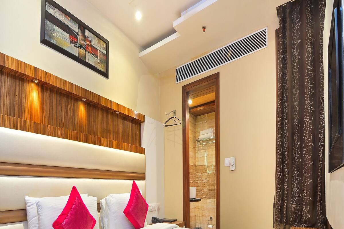 A hotel room with a bed, wooden headboard, a painting, a shower area, and a window with curtains.