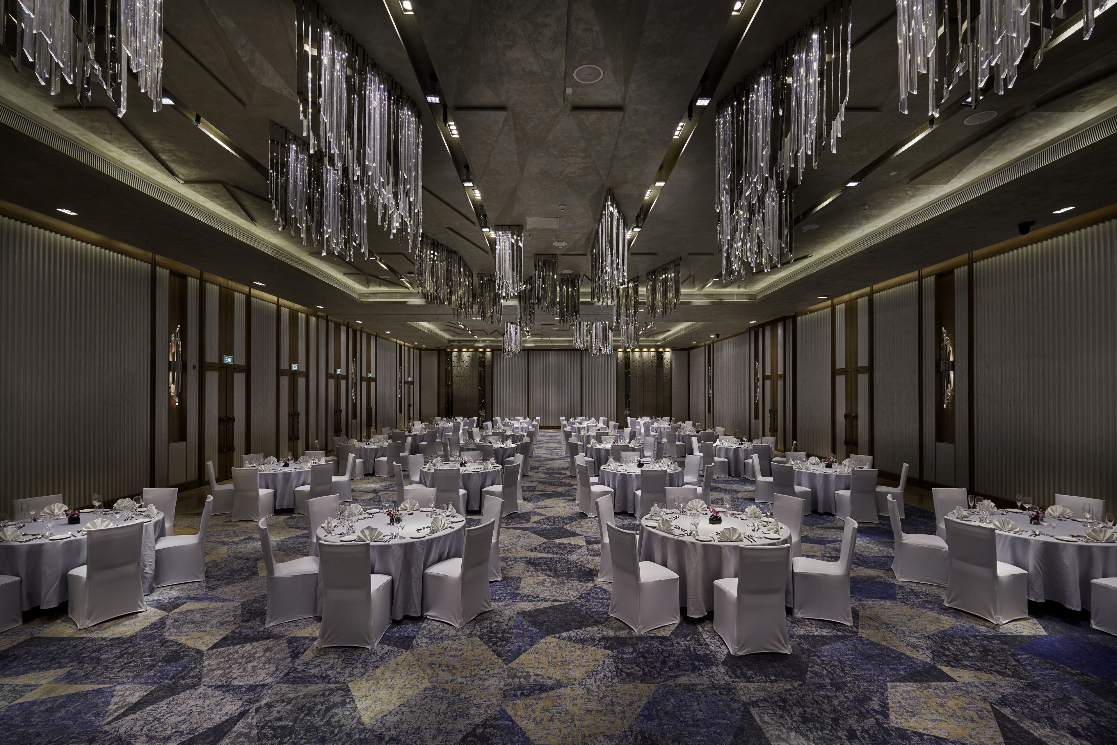 A large banquet hall with round tables and chairs, high ceilings, and hanging chandeliers.