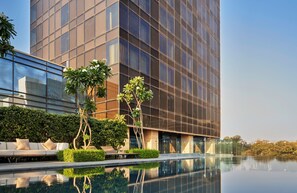 A modern building with a reflective glass facade, a pool with clear water, and a seating area with cushions. - The Den Bengaluru (Bengaluru)