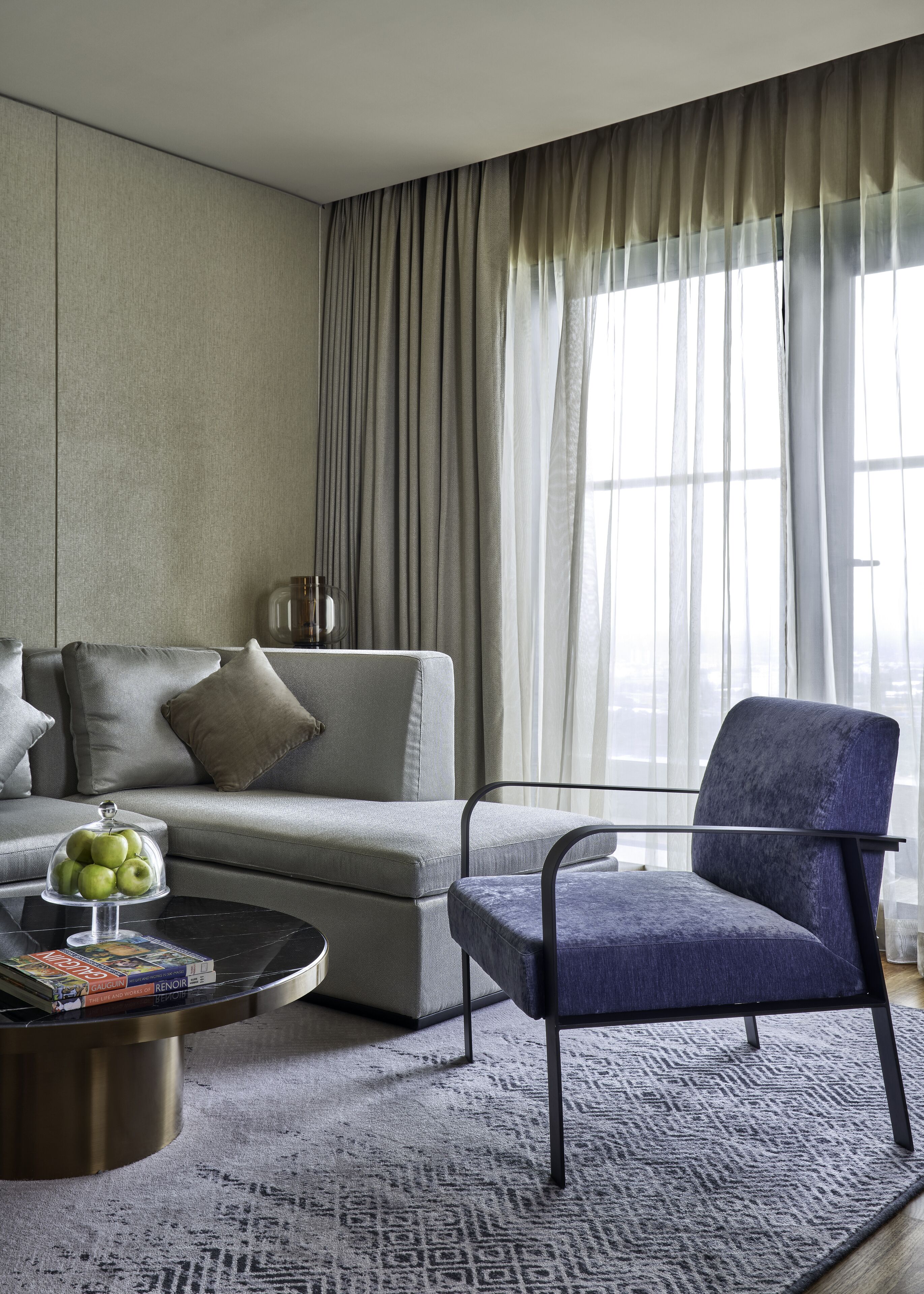 A modern living room with a grey sofa, a blue armchair, a coffee table with a glass dome containing apples, and a patterned rug.