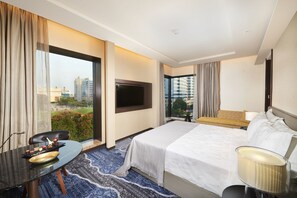 A hotel room with a bed, a desk, a TV, and a view of the cityscape. - The Den Bengaluru (Bengaluru)