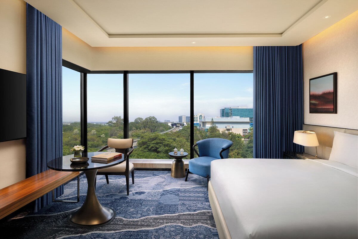 Une chambre d’hôtel avec un grand lit, un bureau avec une chaise, une petite table avec une chaise, une vue sur le paysage urbain à travers de grandes fenêtres et un tableau au mur.