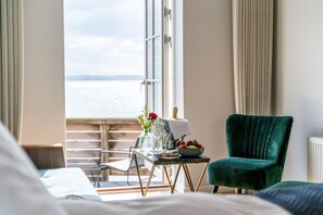 A hotel room with a balcony, a dining table with a bowl of fruit, a green armchair, and a bed with a white comforter. - Kaløvig Badehotel (Aarhus)