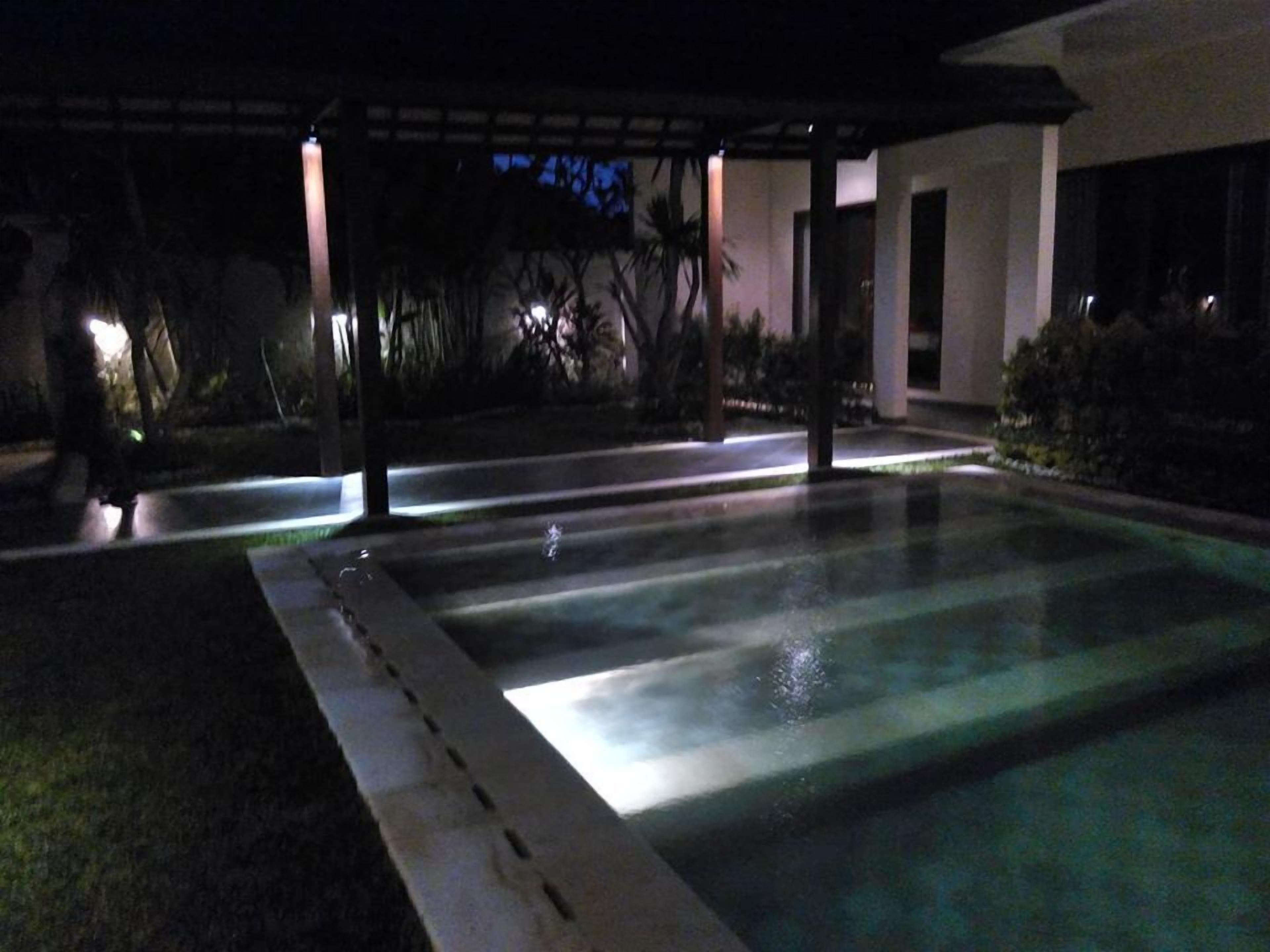 Vista nocturna de una piscina con zona cubierta y un edificio al fondo.